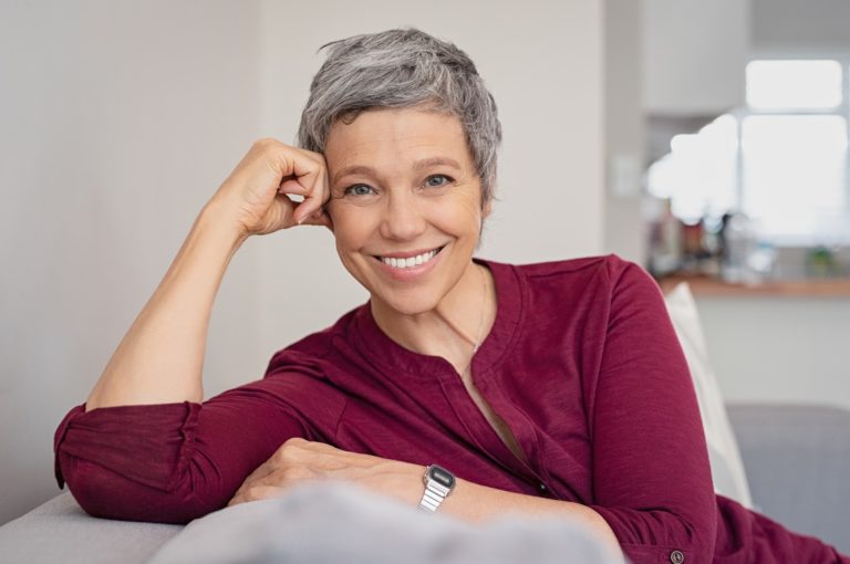 A smiling woman with short gray hair, wearing a maroon top, leaning on her arm and sitting on a couch in a relaxed pose.