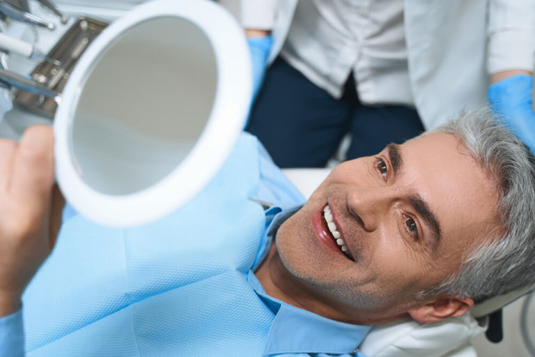 A male dental patient laying back in a dental chair holds a mirror above his head and is smiling as he checks his teeth
