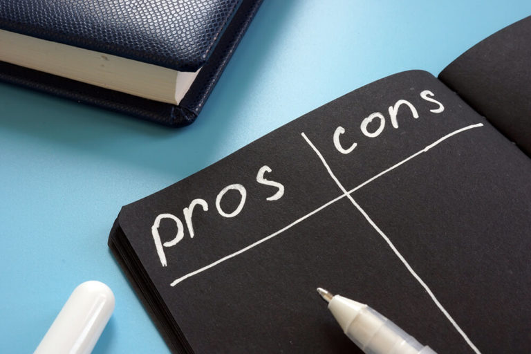 Close-up of a notebook with 'pros' and 'cons' columns written on a black page, next to a white pen, on a blue desk.