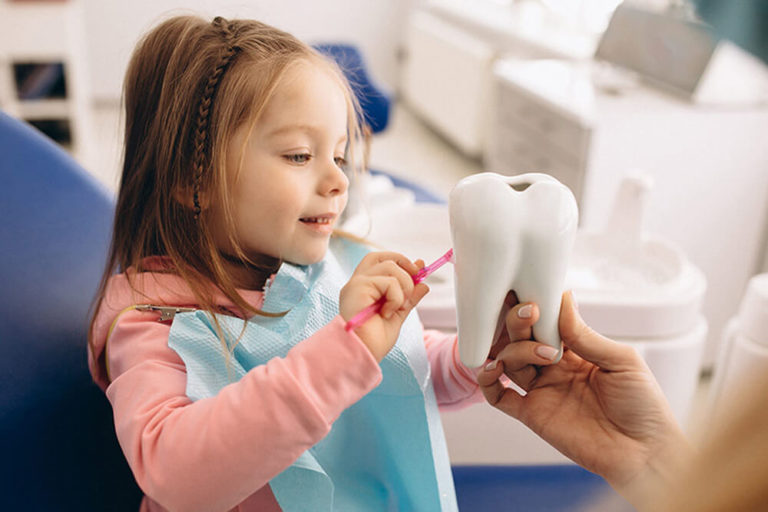 A young girl at a dental office, practicing brushing on a large tooth model held by a dental professional.