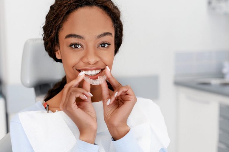 A smiling woman holding a clear dental aligner in front of her teeth, sitting in a dental chair.