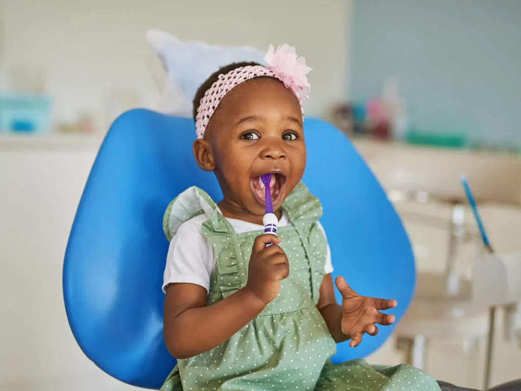 A young child sitting in a dental chair, smiling and holding a purple toothbrush, wearing a green dress and pink headband.