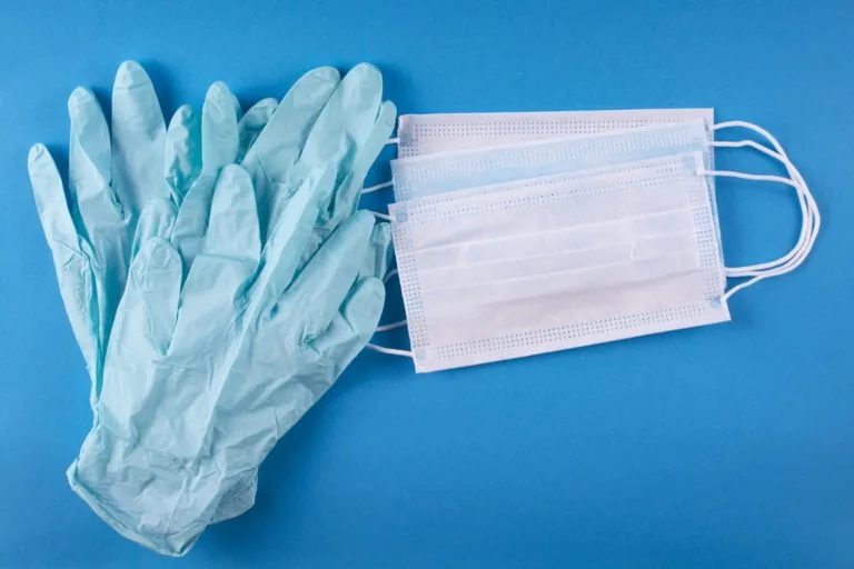 Pair of blue disposable gloves and several white surgical masks placed on a blue background.