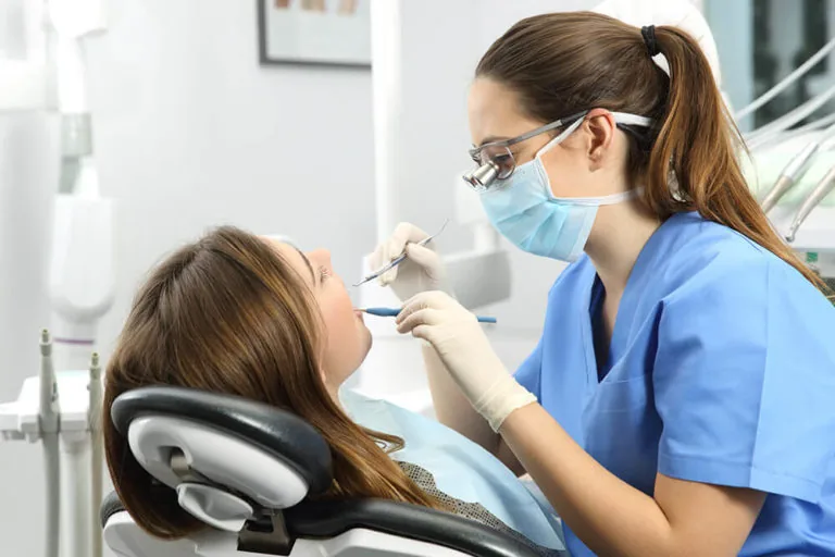 A dental professional wearing a mask and protective eyewear performing a dental check-up on a patient lying in a dental chair.