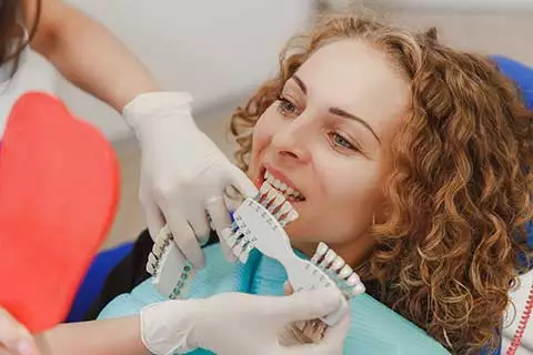 A woman with curly hair at a dental appointment, having her teeth shade matched using a shade guide by a dental professional.