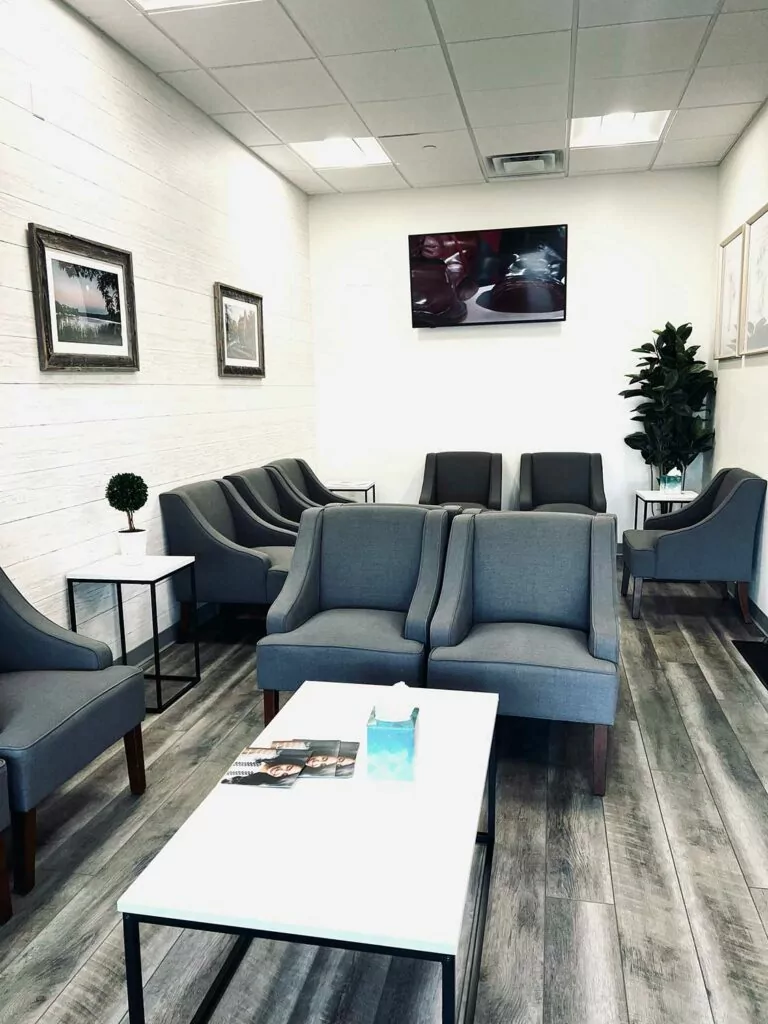 A modern dental office waiting room with gray chairs, a white coffee table, framed pictures on the walls, and a TV mounted above.