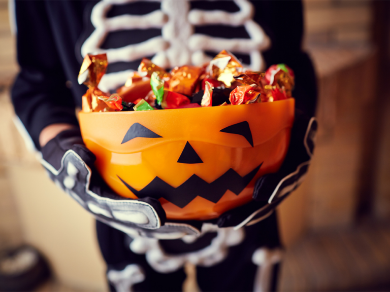 A person in a skeleton costume holding an orange pumpkin-shaped bowl filled with wrapped Halloween candies.