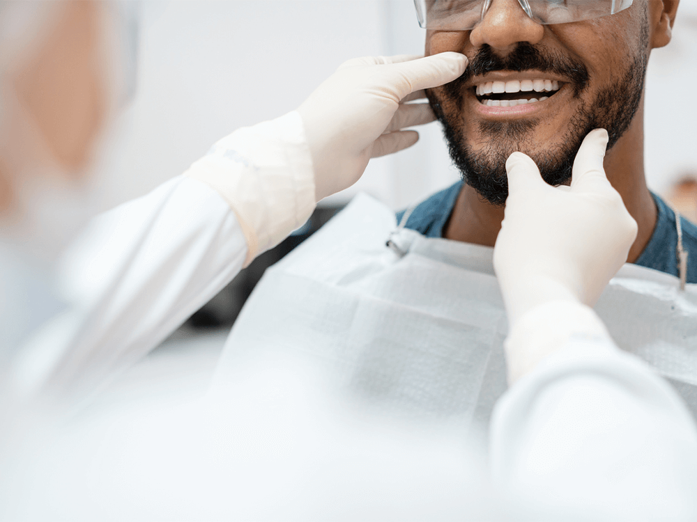 A dentist's hands examining a patient's smile, with the patient wearing a dental bib and protective glasses.