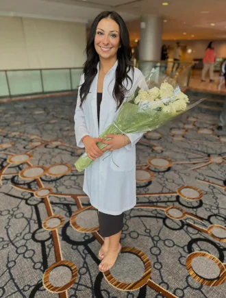 Dr. Efstratia Rigopoulos in white coat holding bouquet of white roses wrapped in green paper, standing barefoot on patterned carpet in formal indoor setting.